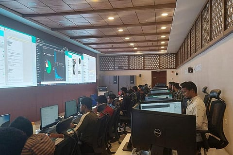People in a control room seated in rows in front of computers with a large screen on the wall ahead of them