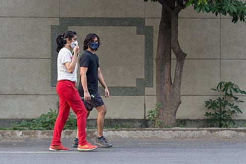 A couple walking on the road, both wearing masks. The man is dressed in black, the woman is wearing red pants.