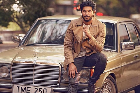 Dulquer Salmaan is striking a stylish pose, sitting on top of a car in the image.