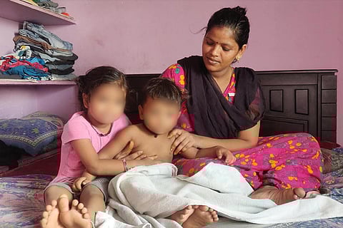 Sathya in a pink nightie wearing a brown shawl across her is sitting on a bed with her two children Sadhana and Eshanth. Eshanth is not wearing a shirt, Sadhana is in a pink dress