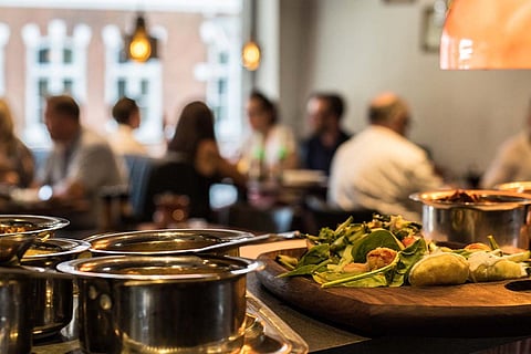 Food kept steel vessels in a restaurant. People seen sitting in the background.