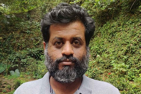 Jeo Baby with salt and pepper beard wearing a grey t shirt and standing against greenery