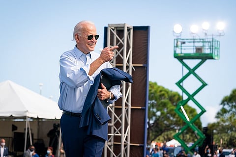 Joe Biden wearing sunglasses has his coat in his left hand and his points to the crowd