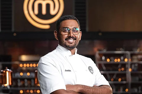 Justin Narayan in a white apron, smiling, in front of the MasterChef logo