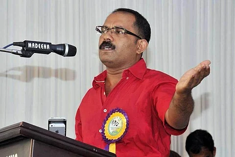 KM Shaji wearing a red shirt and speaking at an event standing