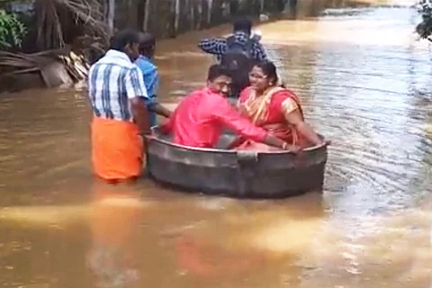 The couple, decked up in pink wedding attires, sit in a copper cooking vessel to travel through a flooded road. Three people are seen steering the vessel through the water.
