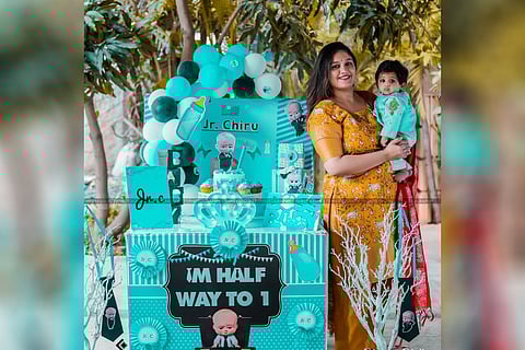 Meghana Raj is seen holding Jr Chiru, who is dressed in blue ethnic clothes. cake, baloons and other decoration items are seen in the background.