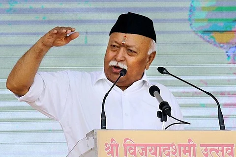 Mohan Bhagwat wearing black cap and white shirt and talking into a mic on a podium