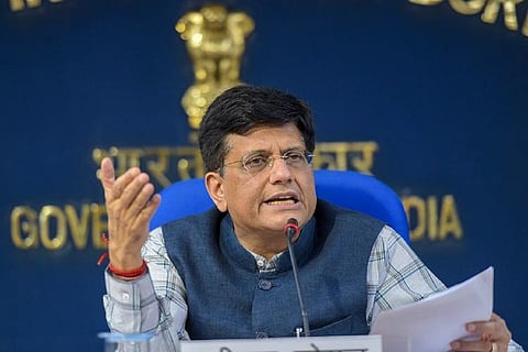 Piyush Goyal in a white shirt with blue check-lines. He is also wearing a blue sleeveless jacket. He is seated on a blue chair and is addressing an audience, with a set of papers in his left hand.