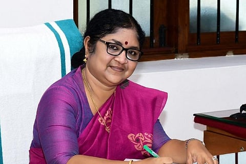 Kerala Higher Education Minister R. Bindu is smiling while seated at her desk, wearing a purple saree and glasses, holding a pen.