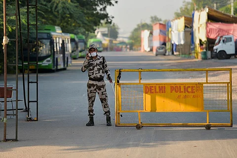 A police personnel speaking on a phone and standing near a yellow-colored Delhi Police barricade, on an empty street