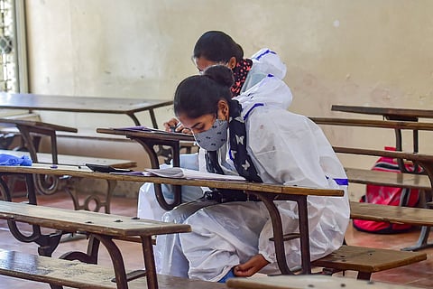 Two students sitting at individual wooden desks in an examination hall wearing PPE and masks
