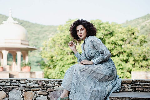 Taapsee Pannu in a grey dress sitting with a monument in the background