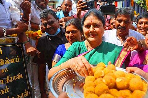 VK Sasikala paying respects at the MGR Memorial, wearing a green saree she can be seen smiling into the camera, a plate of laddoos in front of her