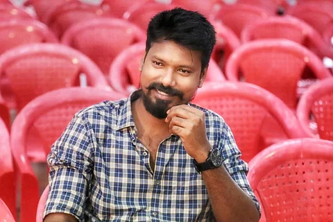 Koozhangal director Vinothraj in a checked shirt and sitting with red chairs behind him
