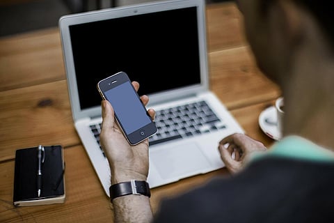 A man sitting at a desk with a laptop in front of him and looking at a phone in his hand