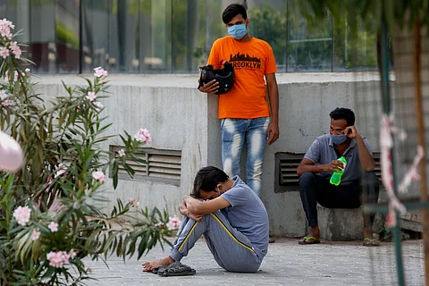 Three young men, one sits with his head bowed, one stands behind in an orange shirt wearing a mask, and a third sits on a platform holding a bottle of water
