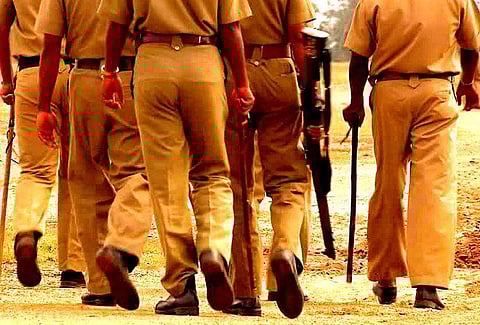 A group of police men walking away from the camera. Their backs, from waist below, face the camera. They are khaki-clad. Some are holding lathis and rifles.