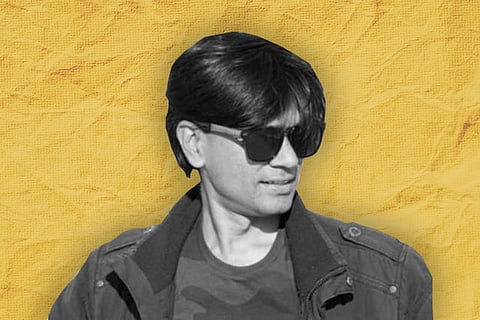 A man with medium-length dark hair wearing sunglasses and a dark jacket over a camouflage t-shirt, looking to the side. The background is a textured yellow.