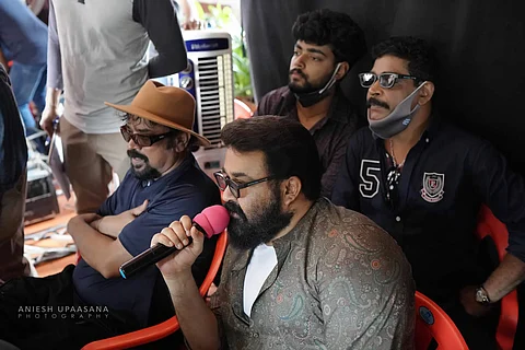 Mohanlal on the first day of shooting of his directorial debut Barroz, seated and speaking into a mic. Seen with him are cinematographer Santosh Sivan and producer Antony Perumbavoor.