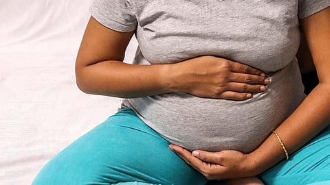 A pregnant woman sitting cross-legged, wearing a grey T-shirt and teal pants, holding her belly with both hands.