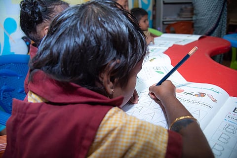 A child writing in a text book with her pencil while sitting at her desk at an anganwadi