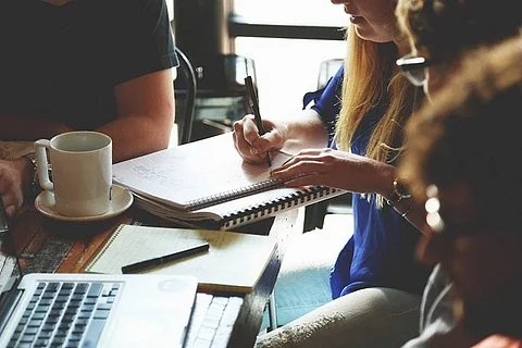 A picture of woman writing something on a book with pen and people sitting around her