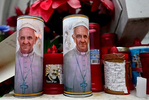 Candles with portraits of Pope Francis and Padre Pio are set at the bottom of a statue of Pope John Paul II outside the Gemelli Hospital where Pope Francis was hospitalized for tests and treatment for bronchitis in Rome, on February 19, 2025. Pope Francis, who has been diagnosed with pneumonia in both lungs, passed a "peaceful night", the Vatican said on 19 February 2025, amid growing concerns over the 88-year-old's condition.