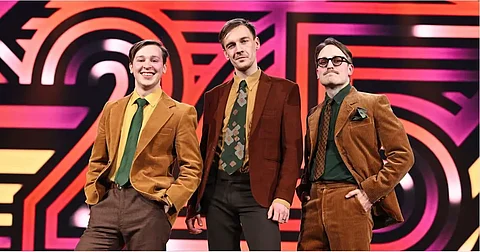 The Finnish comedy trio KAJ pose in retro-style brown and mustard-colored suits against a bold, colorful Eurovision 2025 stage backdrop. The three members smile confidently, exuding quirky charm ahead of their performance of Bara Bada Bastu.