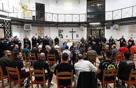 Pope Francis sits in front of an altar inside the rotunda of Regina Coeli Prison, surrounded by seated inmates and uniformed prison staff during a Holy Thursday visit.