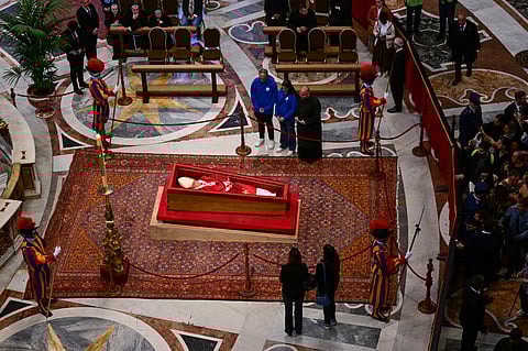Mourners and members of the Swiss Guard gather around the open casket of Pope Francis, lying in state inside a grand church adorned with marble floors and a red carpet, as people pay their final respects.