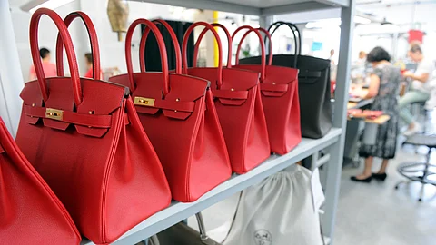 Rows of red Hermès Birkin bags are displayed on a shelf inside the Maroquinerie de la Tardoire, an Hermès workshop specializing in calfskin leather goods, in Montbron, southwestern France. Artisans can be seen working in the background.