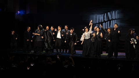 The cast of Liwanag Sa Dilim takes a celebratory curtain call on stage, with the musical’s title projected in the background. Performers are dressed in dark costumes, some raising their arms in triumph and smiling at the applauding audience.