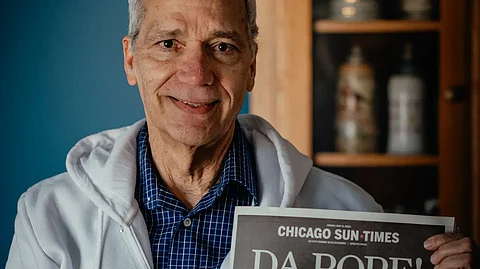John Prevost, wearing a white hoodie and blue checkered shirt, smiles as he holds a Chicago Sun-Times newspaper with the headline “DA POPE!” featuring a photo of his brother, Pope Leo XIV, on the front page.