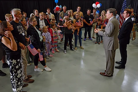 Deputy Secretary of State Christopher Landau and Homeland Security Deputy Secretary Troy Edgar address a group of Afrikaner refugees from South Africa during a welcome ceremony at Atlantic Aviation Dulles in Virginia on 12 May 2025. The group, including children, holds small U.S. flags as they listen.