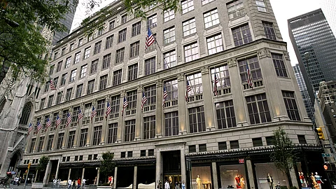 Exterior view of Saks Fifth Avenue in New York City, with American flags lining the historic building's facade. Shoppers are seen walking past display windows at street level.