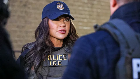 A U.S. Immigration and Customs Enforcement (ICE) officer wearing a cap and tactical vest labeled “POLICE ICE” speaks to colleagues during an operation.
