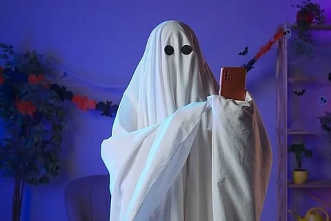 CREEPING INTO YOUR DMs — A person in a classic white ghost costume scrolls through their phone, capturing the eerie, playful spirit of ghostlighting in modern dating.
