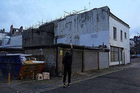 People take photographs of a graffiti street art by street artist Banksy, in Bayswater, west London on December 22, 2025. British street artist Banksy unveiled his latest work in central London with typical mystery amidst speculation over the origin of an identical work which appeared on the same day. The black and white mural painted on the side of an old building shows two people in winter hats and wellington boots appearing to be lying down looking up in the direction of the sky.