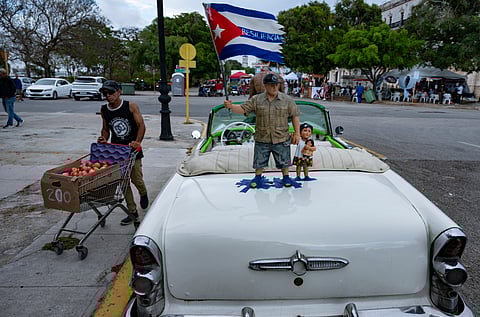 An apple seller walks past a car and a figure holding the Cuban flag with the word ‘Resilience’ after a blackout in Havana.