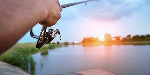 Fisherman holds spinning reel in hands and cathes fish in lake or river at sunset at summer day