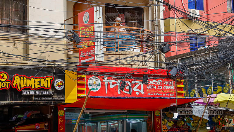 A woman stands on a balcony obscured by power cables and restaurant signboards at a building in Dhaka’s Banasree.