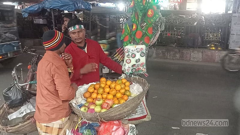 A fruit vendor wearing a band to mark Victory Day speaks to a rickshaw driver selling national flags on the street in Rangpur on Tuesday, Dec 16, 2024. Credit: Samiha Mehbub Hiya