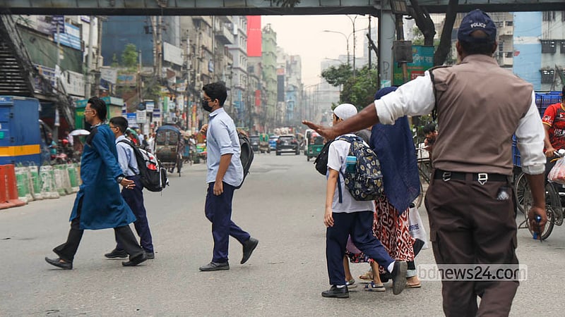 Despite the footbridge at Malibagh intersection, people often cross underneath, forcing traffic police to stop vehicles to prevent accidents. A traffic police officer raises a hand to halt traffic and helps students and parents cross safely. Photo: Shamsul Haque Ripon