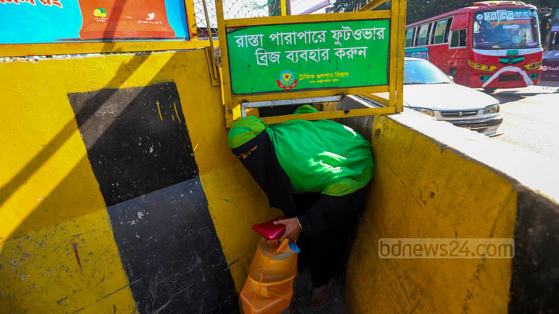 ঢাকার মহাখালীতে সড়ক বিভাজকের উপরে দেওয়া লোহার বেষ্টনীর নিচে কিছুটা ফাঁকা জায়গা পেয়ে ওই পথেই পার হওয়ার চেষ্টায় এক নারী। অথচ কয়েক গজ দূরেই রয়েছে ফুটব্রিজ।