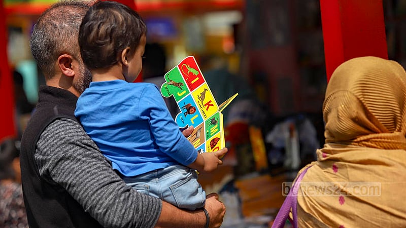 বইমেলায় বাবার কোলে বসেই বর্ণমালার বই পড়ছিল শিশুটি।&amp;nbsp;ছবি: মাহমুদ জামান অভি