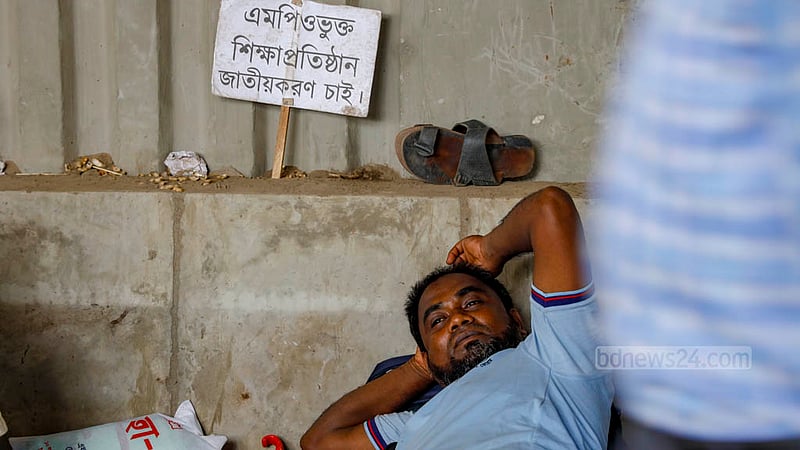 জাতীয় প্রেস ক্লাবের সামনের সড়কের উপরে শুয়ে আছেন এক শিক্ষক, পাশে প্ল্যাকার্ডে লেখা- ‘এমপিওভুক্ত শিক্ষা প্রতিষ্ঠান জাতীয়করণ চাই’।&amp;nbsp;ছবি: মাহমুদ জামান অভি