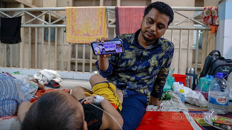 মোবাইল ফোনে কার্টুন দেখিয়ে সন্তান মোহাম্মদ আব্দুল্লাহকে শান্তু রাখার চেষ্টায় বাবা মুক্তার হোসেন। পাঁচ দিন ধরে ডেঙ্গু আক্রান্ত হয়ে একই হাসপাতালে ভর্তি আছেন তারা।&amp;nbsp;ছবি: মাহমুদ জামান অভি