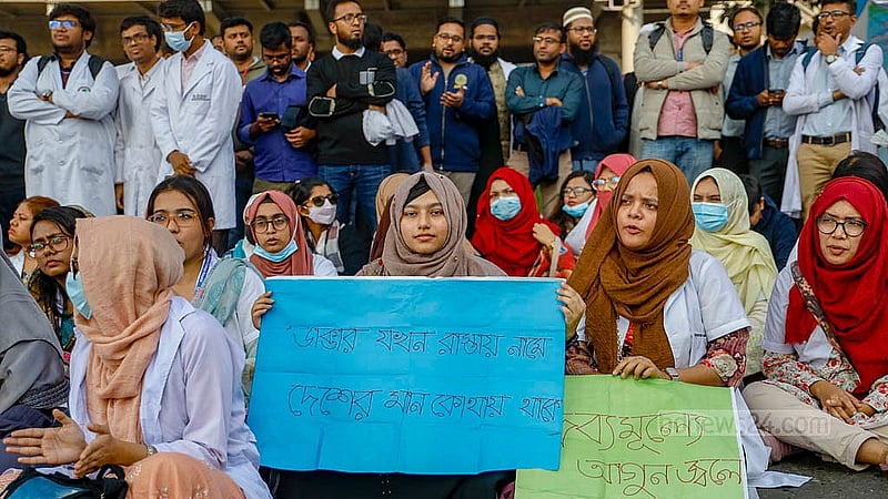 প্ল্যাকার্ড হাতে রোববার শাহবাগ মোড় অবরোধ করে সড়কে বসে আছেন পোস্ট গ্র্যাজুয়েট ট্রেইনি চিকিৎসকরা
