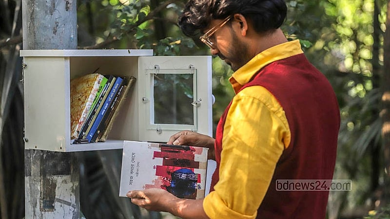 ধানমন্ডি লেকে রাখা কাঠ ও কাচ দিয়ে তৈরি এ বুককেস থেকে বই দেখছেন একজন